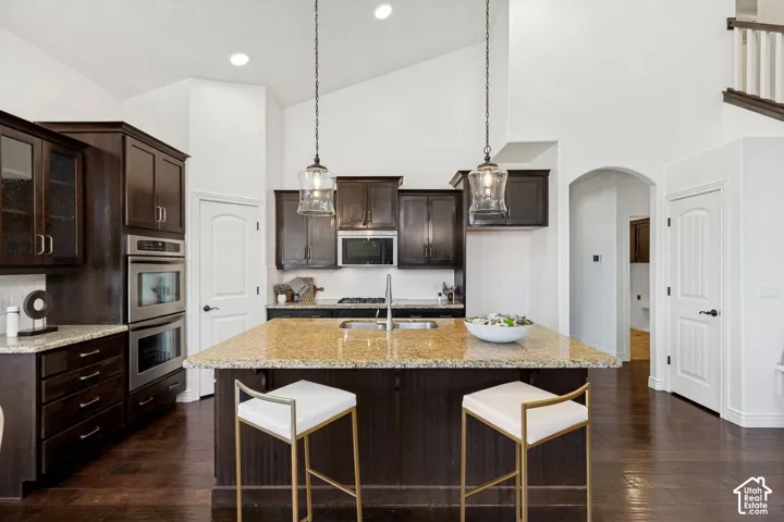 Kitchen featuring dark wood cabinetry, stone countertops, stainless steel double wall ovens, a central island with an integrated sink, and dual glass pendant light fixtures