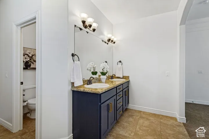 Dual-sink vanity with stone countertop, dark cabinetry, ceramic tile flooring, and wall-mounted light fixtures