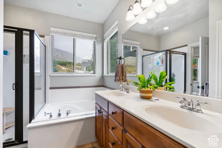 Double vanity featuring light-toned stone countertops, wooden cabinetry, a large wall-mounted mirror, soaking tub, and a glass-enclosed shower
