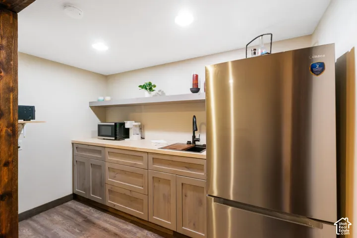 Kitchenette featuring custom wood cabinetry, light-toned countertop, inset sink with matte black faucet, open shelving, and stainless steel refrigerator
