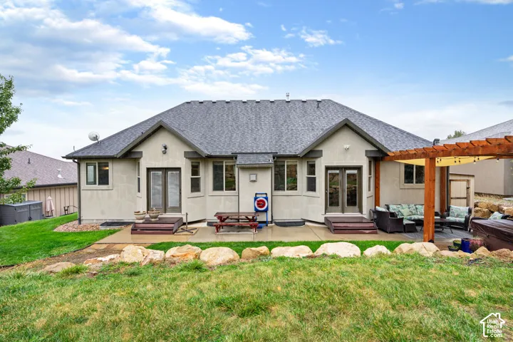 Rear exterior featuring a concrete patio, wood pergola, natural stone landscape edging, dual sets of French doors, and a neutral stucco facade