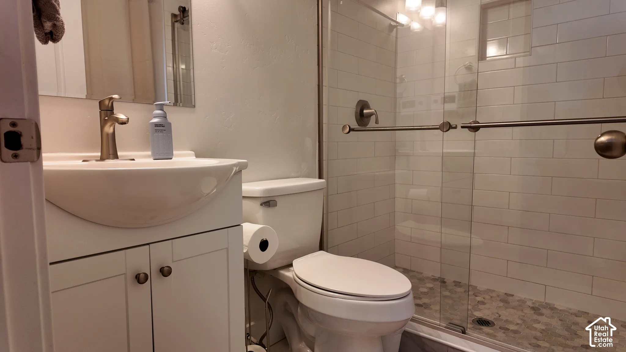 Bathroom featuring a white vanity cabinet with a curved integrated sink, satin nickel faucet, glass-enclosed shower with subway tile surround, and decorative stone-finish floor