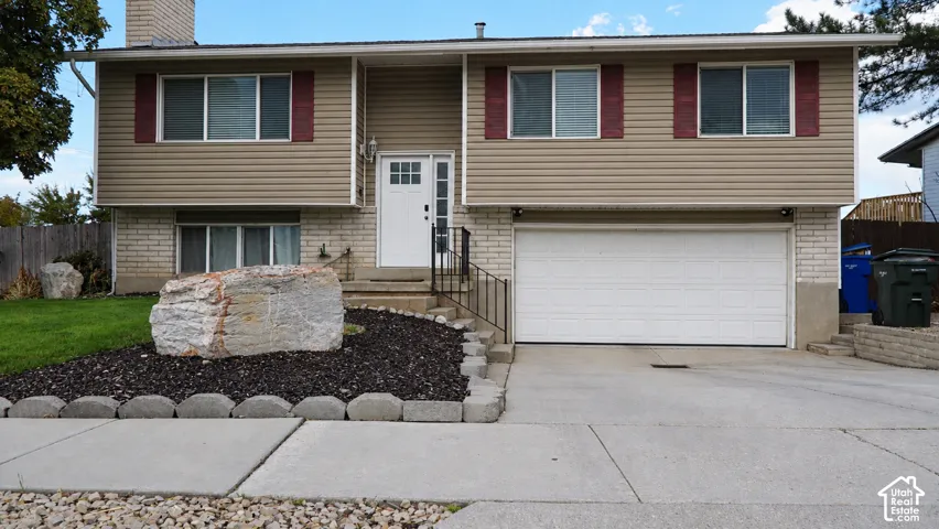 Split-level home featuring a two-car attached garage, stone accent landscaping with mature plantings, concrete driveway and walkway, metal entry railings, and tan vinyl siding with burgundy trim and multi-pane windows across the front elevation