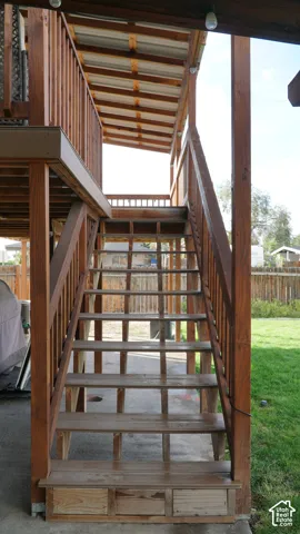 Covered wooden staircase featuring an elevated deck with wood-finish railings and a concrete patio foundation