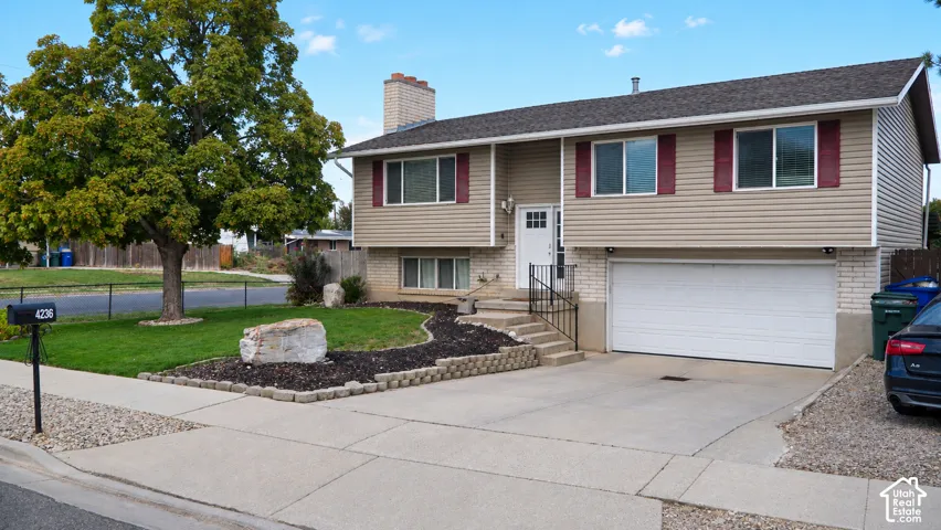 Split-level exterior featuring light-toned siding, brick masonry detailing, an attached two-car garage, concrete driveway, and xeriscaped lawn area with decorative landscape rock