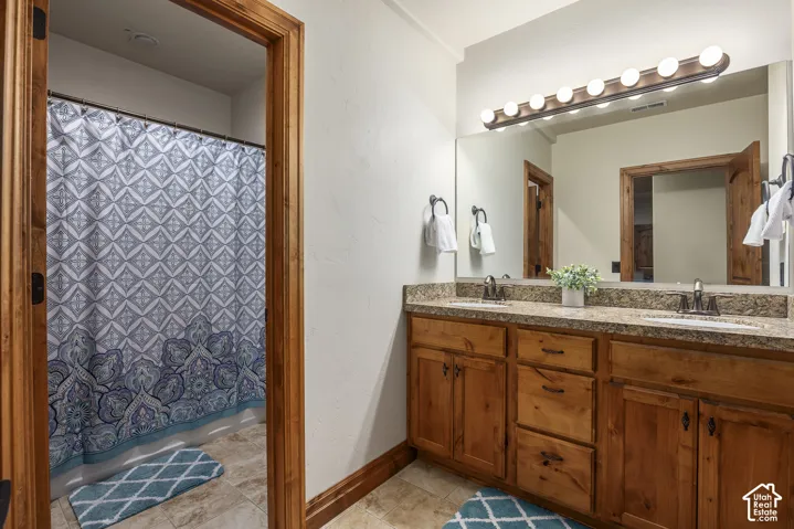 Bathroom featuring a dual-sink vanity with stone-surface countertop, wood cabinetry, large vanity mirror with multi-bulb light fixture, and tile flooring
