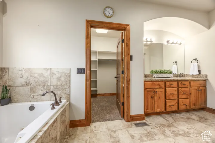 Bathroom featuring a soaking tub with tile surround, wood-finish cabinetry with dual sinks, stone-finish countertops, and an archway entry to a walk-in closet