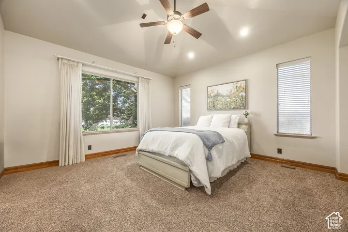 Vaulted ceiling with a central multi-blade ceiling fan, neutral wall-to-wall carpeting, wood-finished trim, and large windows providing natural light