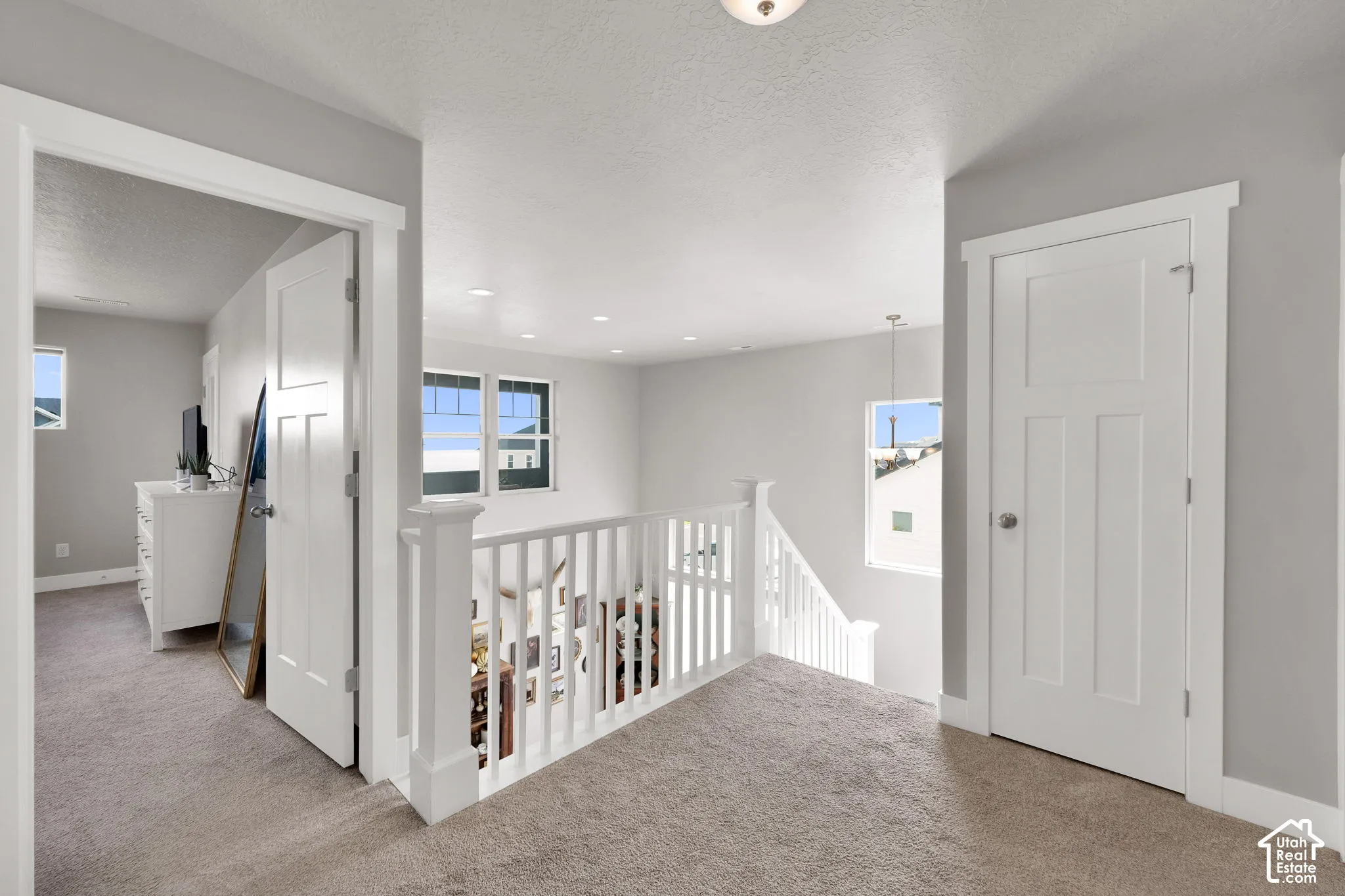 Neutral-toned carpeted hallway featuring a white wooden balustrade, recessed ceiling lighting, and solid-core interior doors with shaker-style detailing