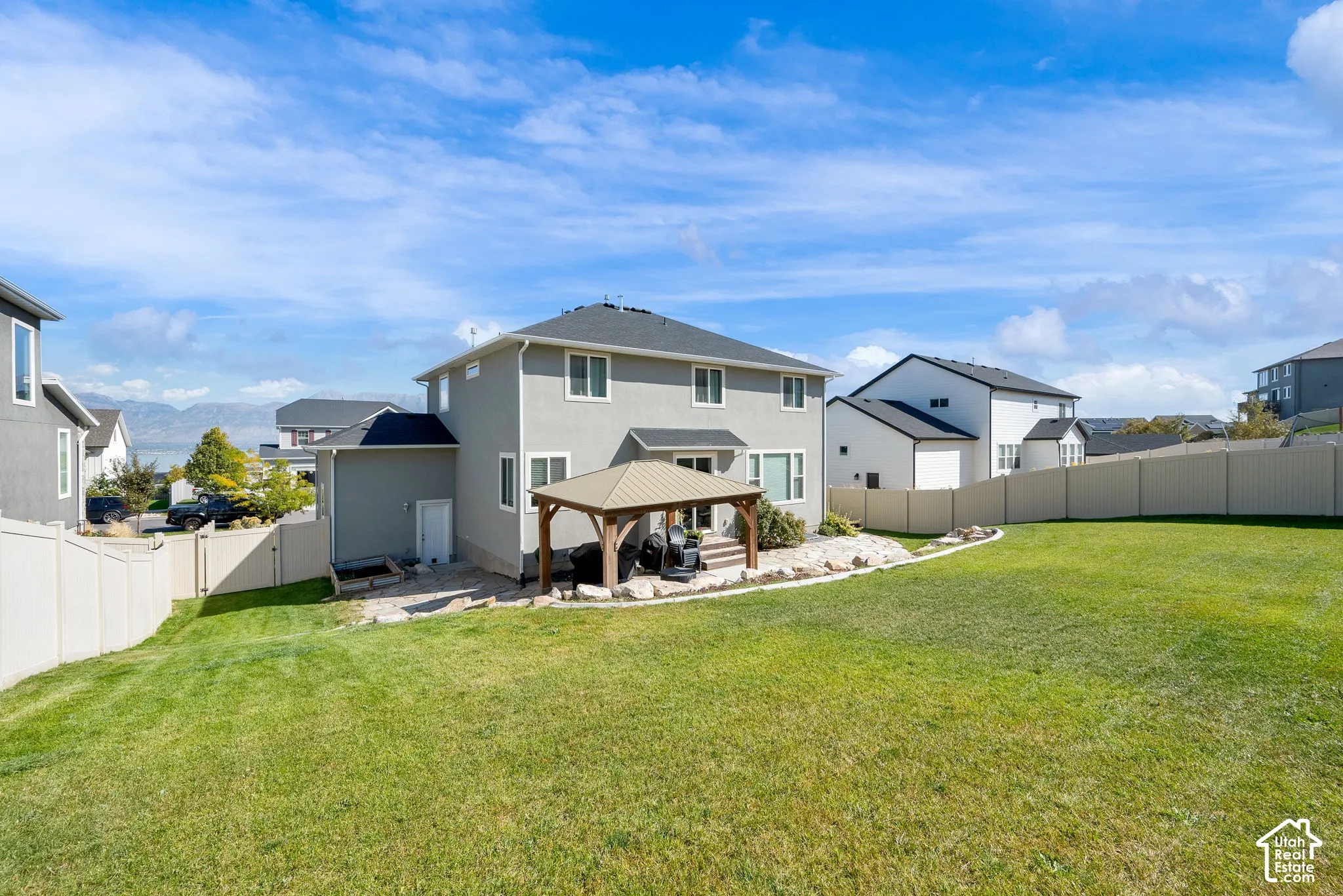 Expansive backyard featuring a manicured lawn, stone-accented patio, permanent gazebo structure, and perimeter vinyl fencing