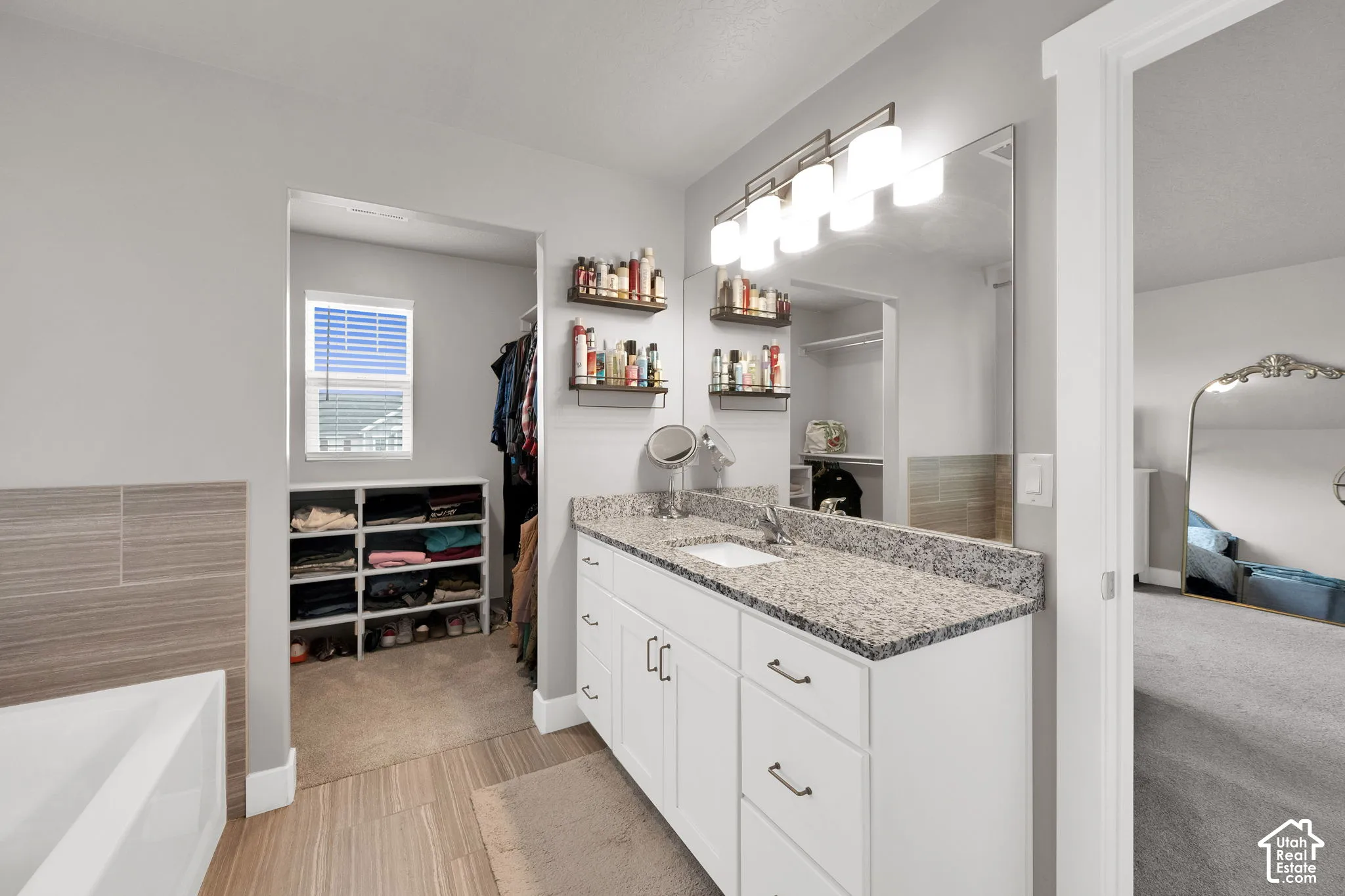 Bathroom featuring a dual-sink vanity with stone-finish countertop, white cabinetry with dark hardware, large wall-mounted mirror, and multi-light fixture, accompanied by a tub with neutral tile surround