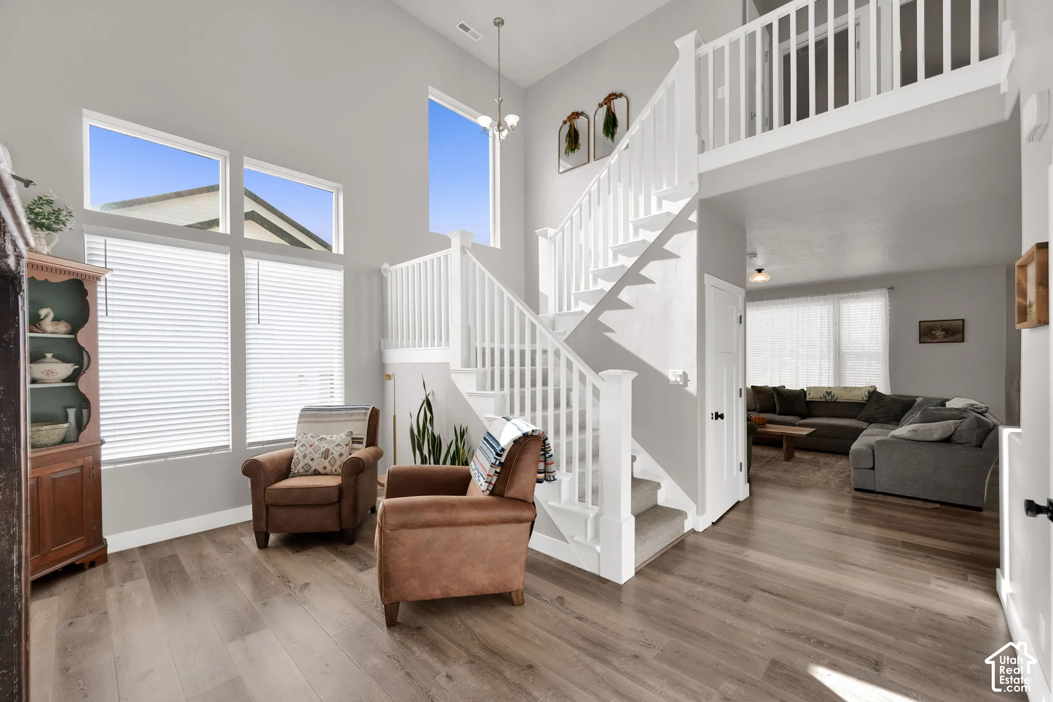 Double-height ceiling, wood-finish flooring, white staircase with balustrade, expansive multi-pane windows, and open upper-level mezzanine