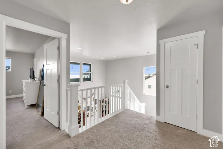 Neutral-toned carpeted hallway featuring a white wooden balustrade, recessed ceiling lighting, and solid-core interior doors with shaker-style detailing
