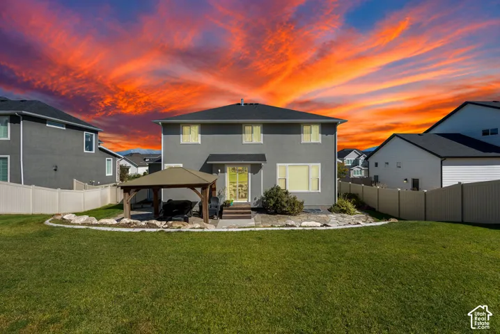 Rear exterior featuring a two-story stucco facade, a permanent hardscape patio, a covered pavilion, and a fenced-in grassy yard
