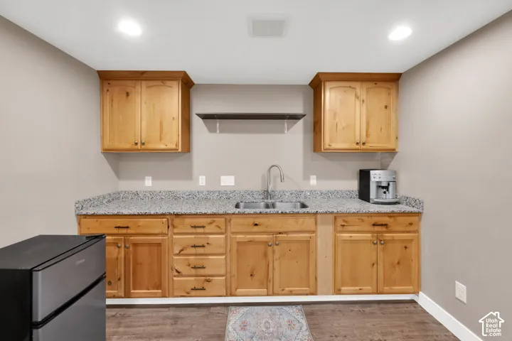 Natural wood-finish cabinetry, stone-surface countertops, dual-basin stainless steel sink with arch faucet, mounted floating shelving, and wood-finish flooring
