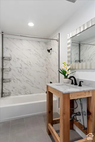 Bathroom featuring a custom wood-framed vanity with a stone countertop, an integrated rectangular sink, matte black widespread faucet, and a shower-tub surround with large-format patterned tile
