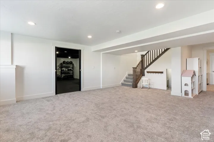 Interior space featuring neutral wall-to-wall carpeting, recessed ceiling lighting, staircase with a stained wood handrail, and a doorway revealing a separate area with dark finished ceiling and flooring