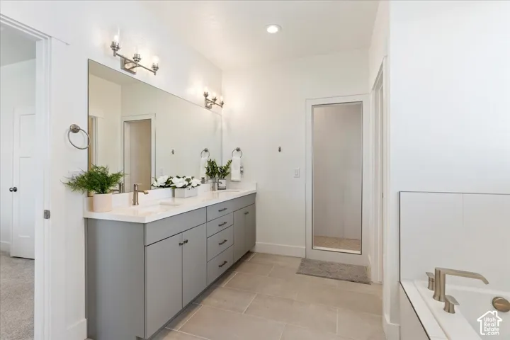 Dual-sink vanity with grey cabinetry, light-toned countertop, large wall-mounted mirror, recessed lighting, and built-in bathtub with a deck-mounted faucet