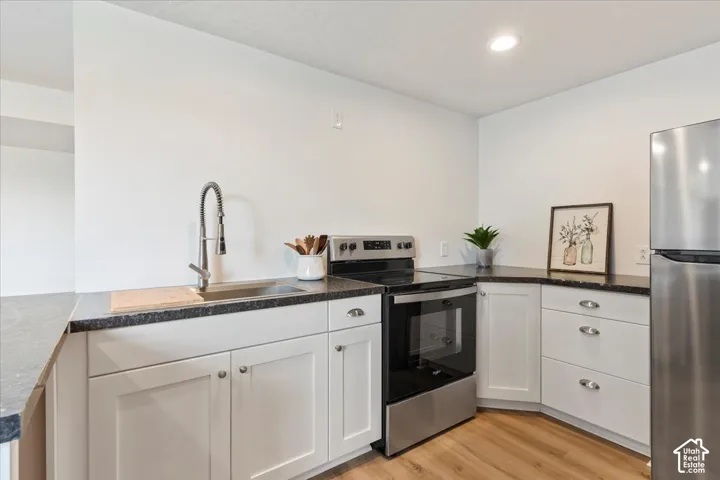 Kitchen featuring white Shaker-style cabinetry, dark countertops, stainless steel appliances, a pull-down sprayer faucet, and wood-finish flooring