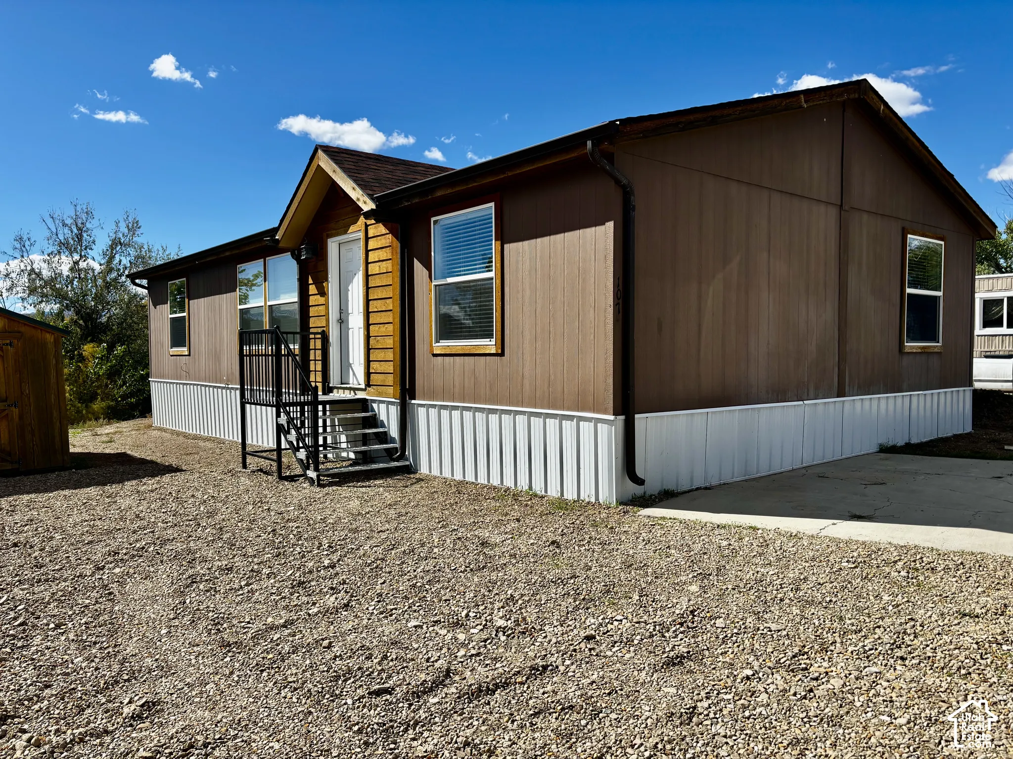 Exterior featuring brown panel siding, wood-accented entryway, metal skirting, shingled gable roof, and concrete pad