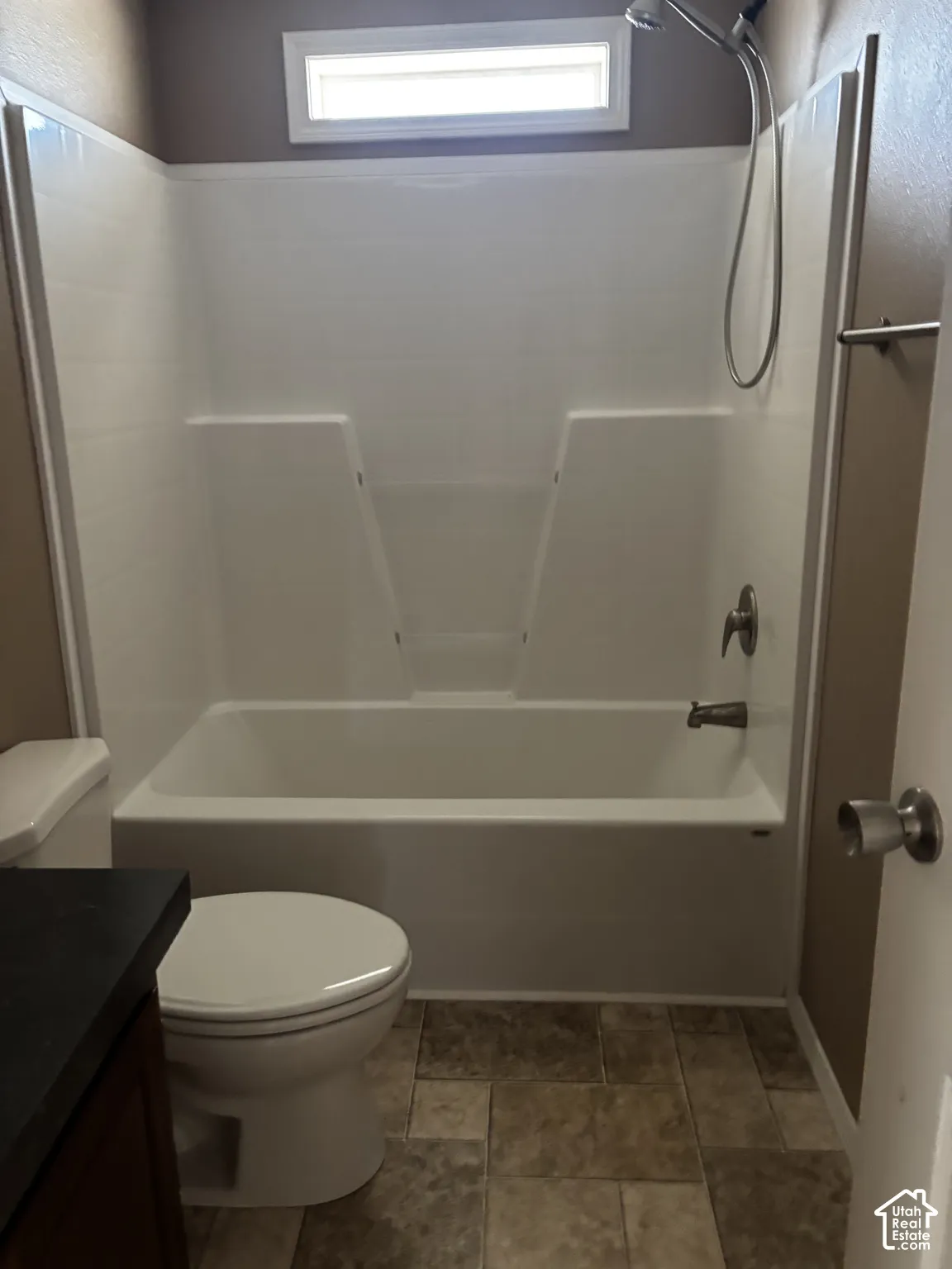 Bathroom featuring a white tub-shower combination with a hand-held showerhead, transom window, and stone-finish tile flooring