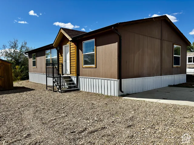 Exterior featuring brown panel siding, wood-accented entryway, metal skirting, shingled gable roof, and concrete pad