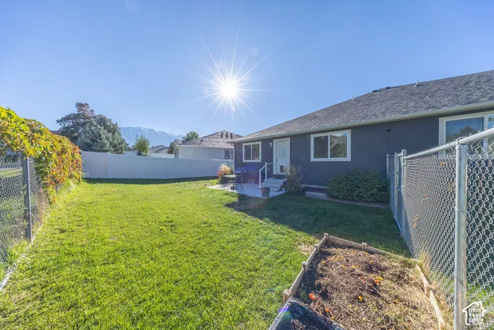 Backyard featuring a grass lawn, concrete patio, raised garden bed, chain-link perimeter fencing, and mountain scenery