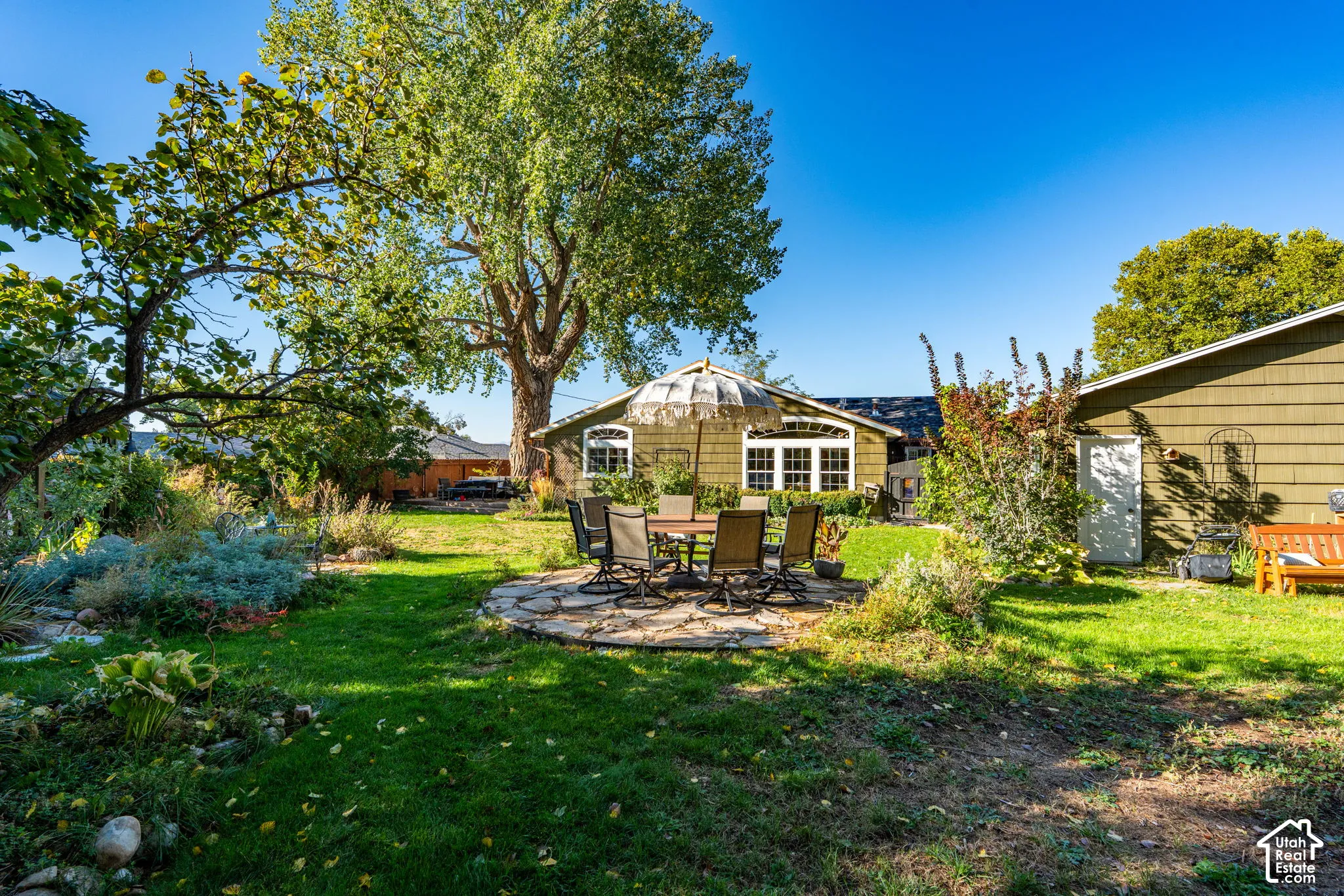 Expansive backyard featuring a natural stone patio, mature trees, landscaped garden beds, and shingle-style exterior siding