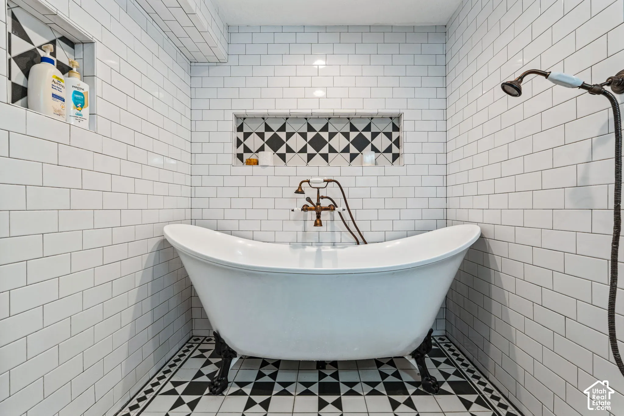 Clawfoot soaking tub with metallic wall-mounted fixtures, white subway tile surround, integrated geometric-patterned tile wall niches, and monochrome patterned floor tile