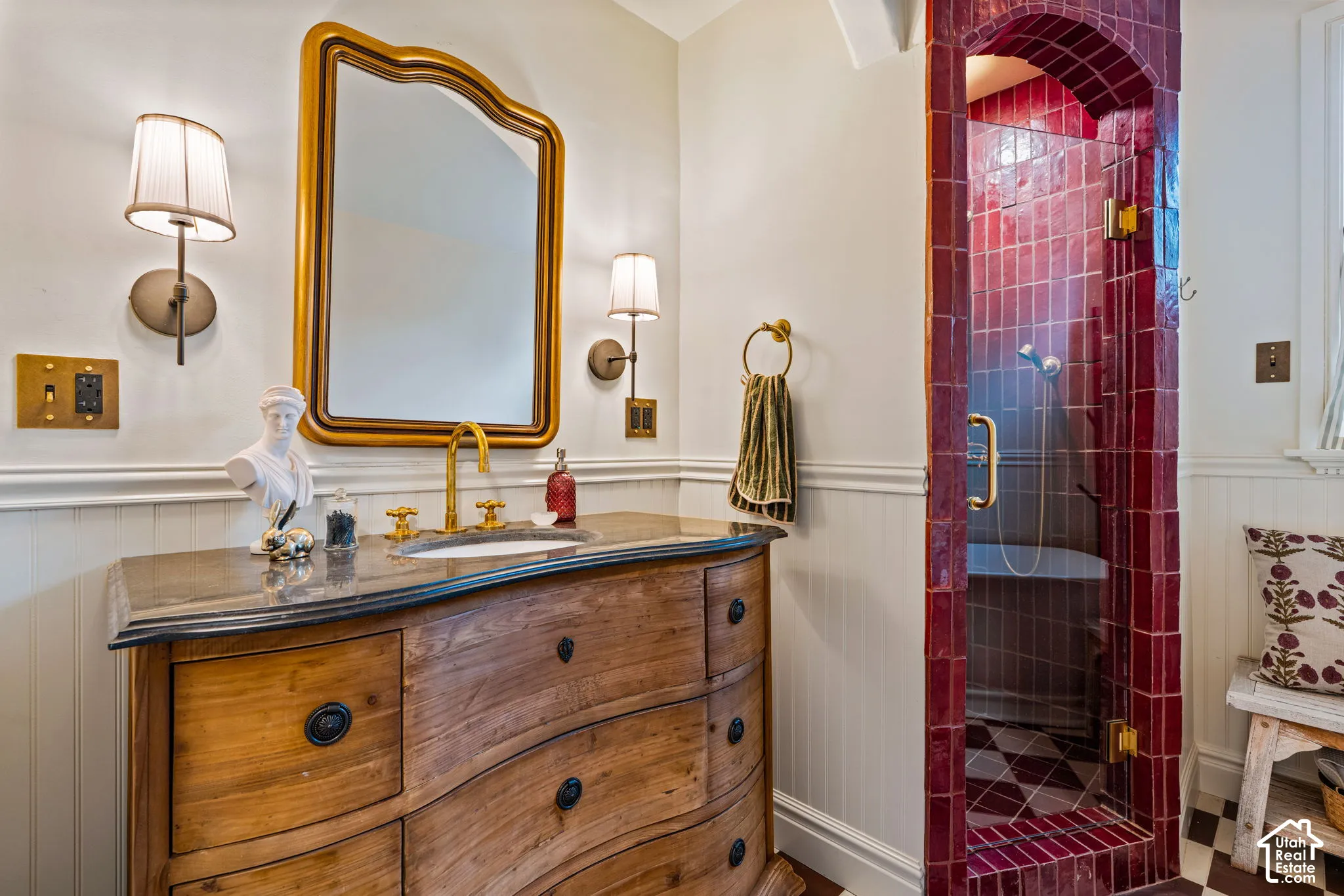 Wood-finish vanity with dark stone countertop, brass faucet, arched shower enclosure with deep-hued glazed tile, and wall-mounted light fixtures