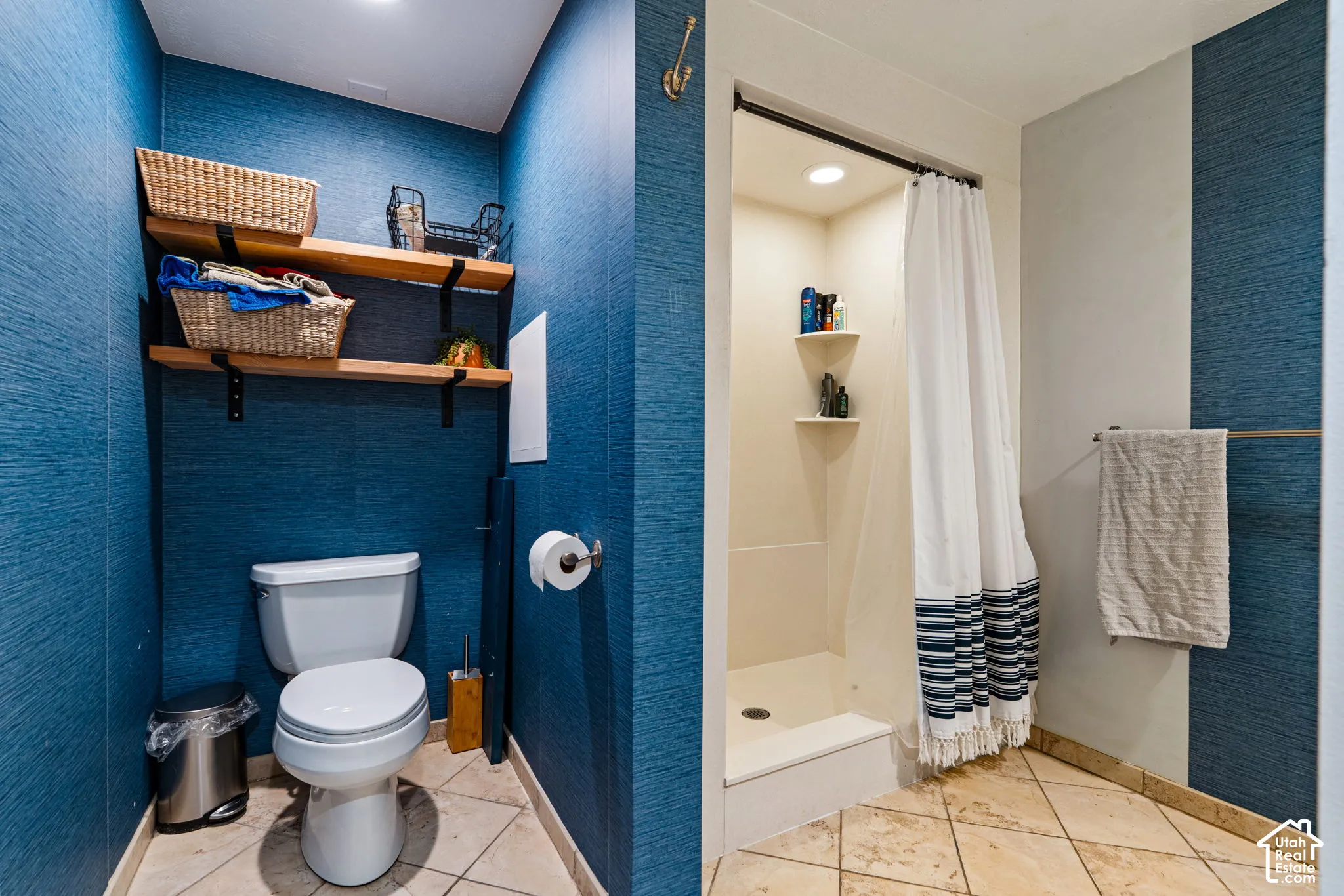 Textured blue wall covering, tiled flooring, built-in wooden shelving, and a tiled shower stall with integrated corner shelving