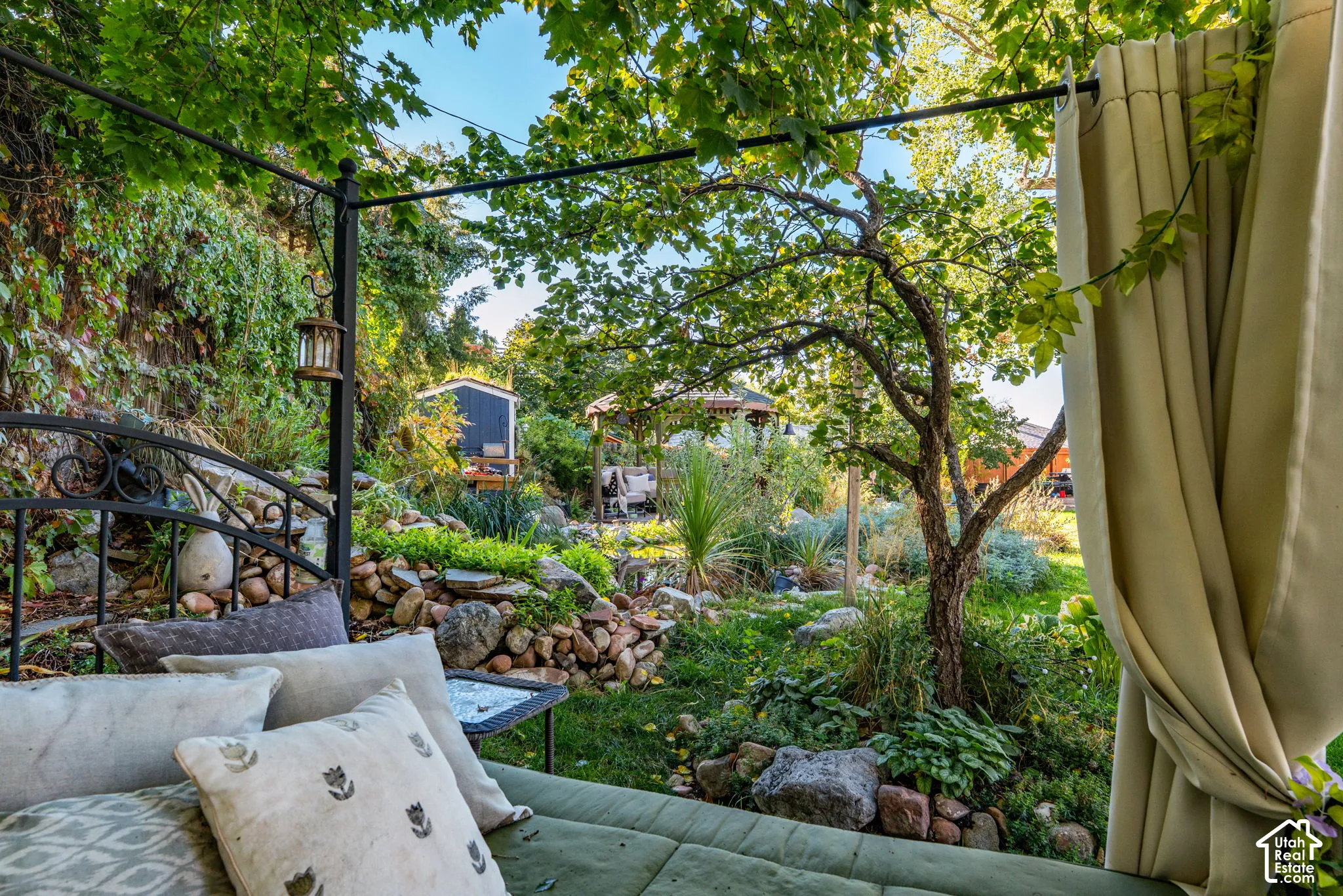 Backyard featuring a tiered stone retaining wall, mature deciduous trees, dense foliage, and a detached wooden structure