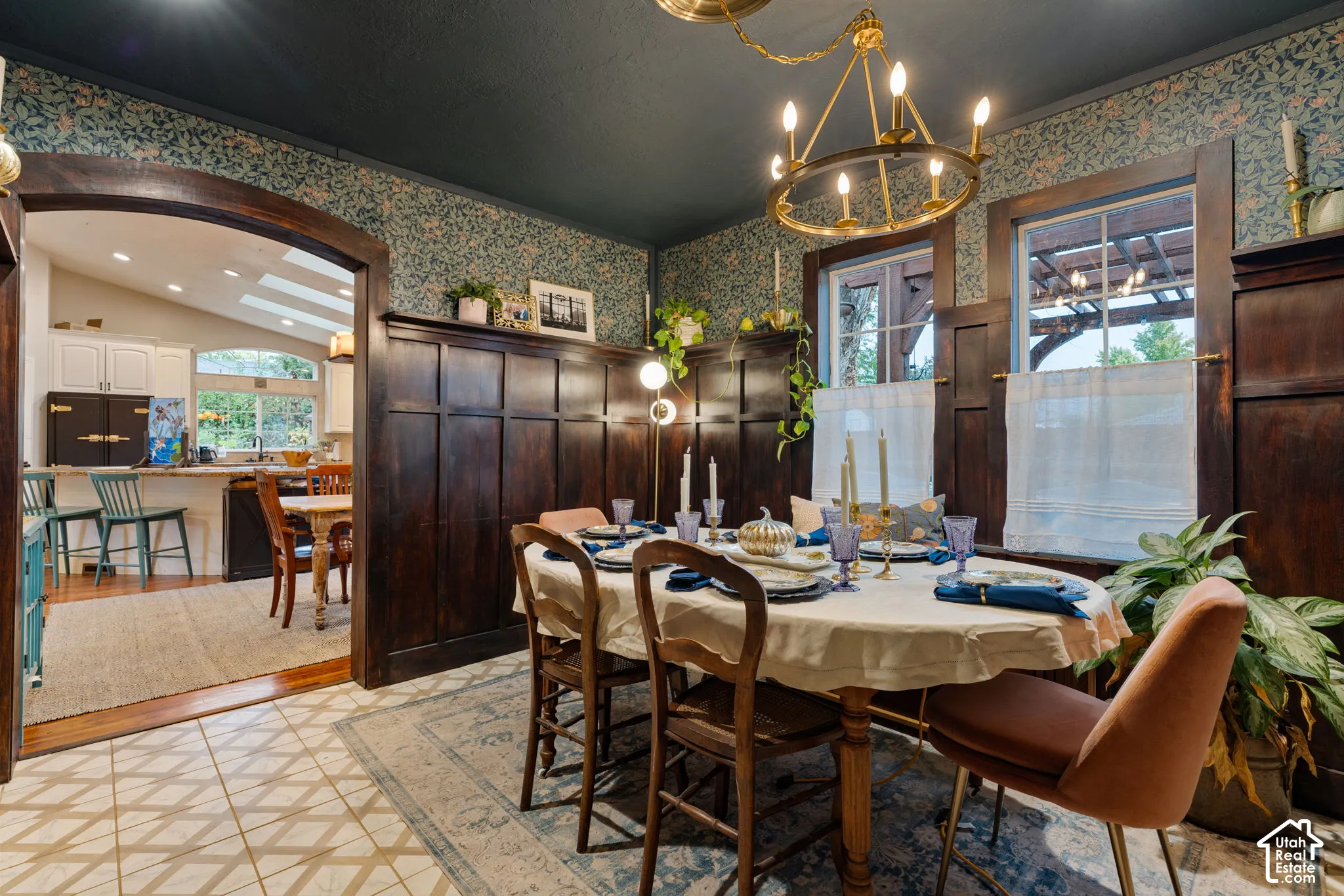 Dining area featuring floor-to-ceiling dark wood wainscoting, patterned wallpaper, a central wagon wheel chandelier, an arched entryway, and tile flooring