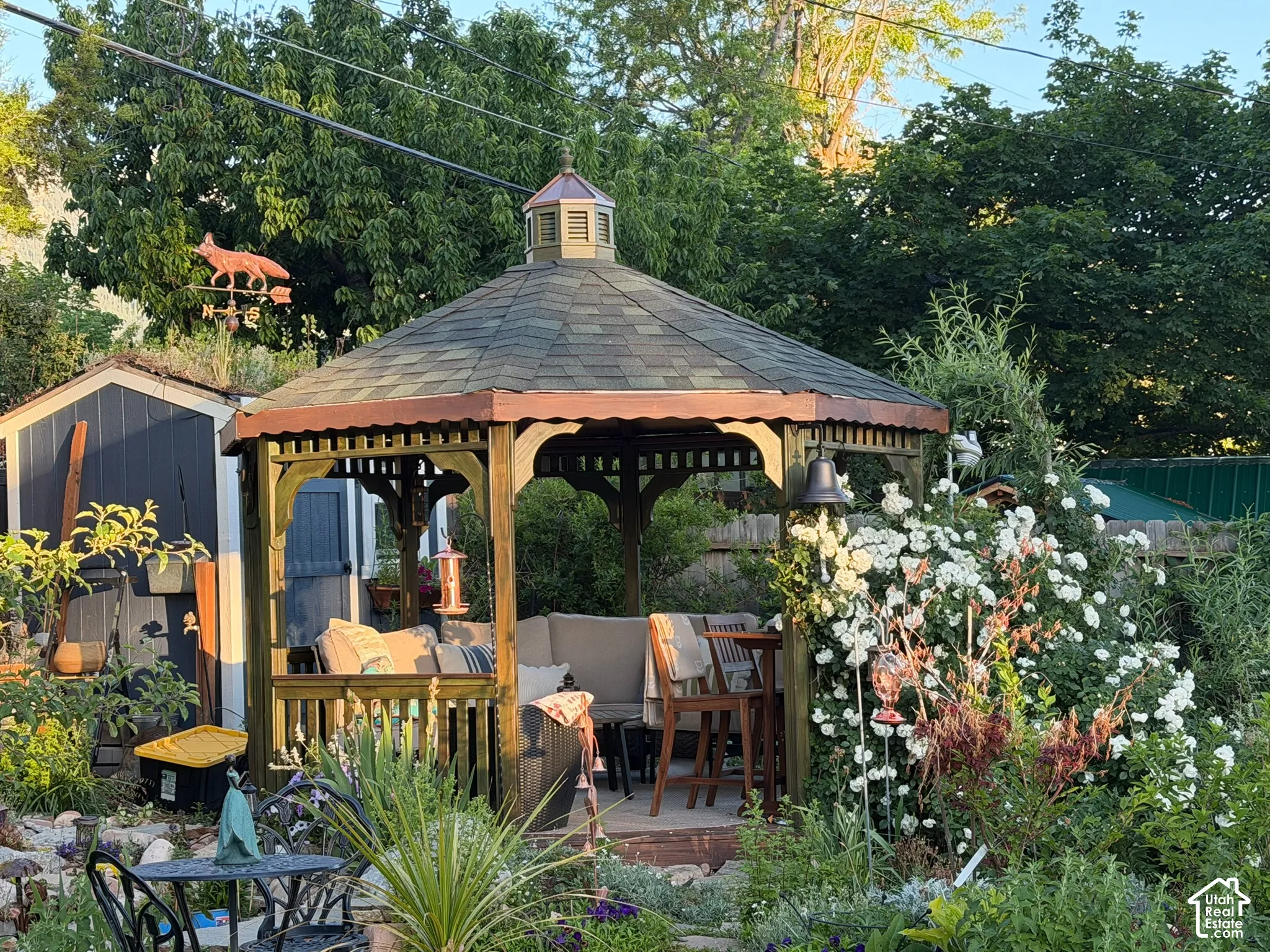 Hexagonal wood gazebo featuring an architectural shingle roof, integrated cupola, and decorative lattice detailing, complemented by landscaped garden beds