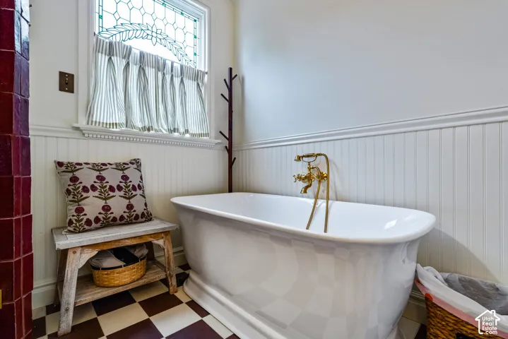 Freestanding white soaking tub with brass deck-mounted telephone faucet, beadboard wainscoting, stained-glass window accent, and checkerboard tile flooring