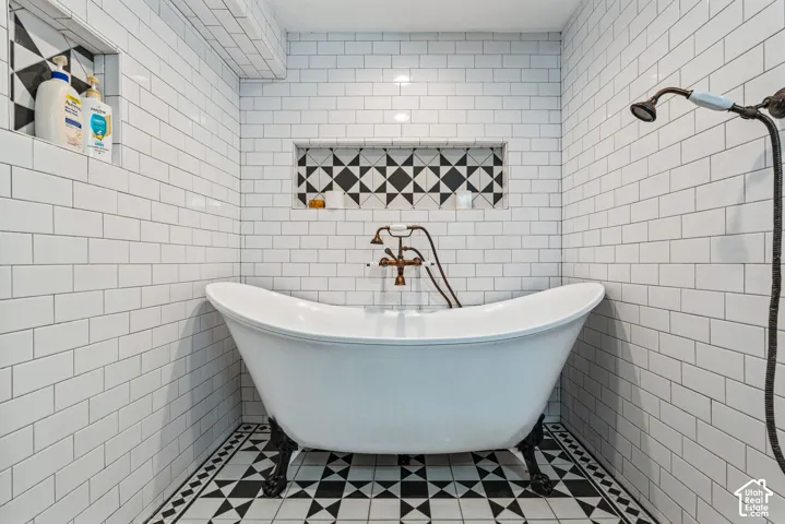 Clawfoot soaking tub with metallic wall-mounted fixtures, white subway tile surround, integrated geometric-patterned tile wall niches, and monochrome patterned floor tile
