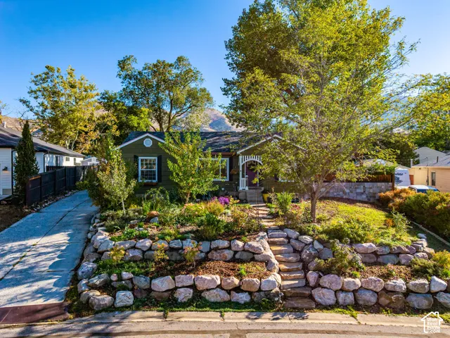 Single-story residence featuring green exterior siding, tiered rock retaining walls, custom stone steps, mature landscaping, and mountain scenery