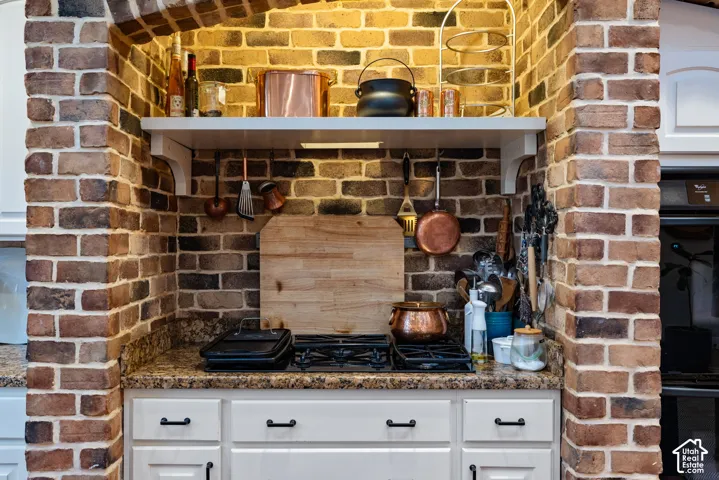 Exposed brick archway with a built-in recessed cooktop nook, white cabinetry, stone-finish countertops, and a mounted wooden display shelf