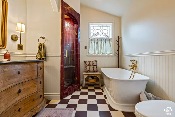 Freestanding clawfoot tub with brass-finish faucet, custom-tiled arched shower entry, checkered tile flooring, wainscoting, and decorative leaded glass window
