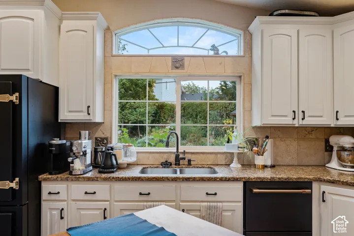 Kitchen featuring white shaker-style cabinetry, stone tile backsplash, stone countertop, dual-basin sink with a matte black faucet, and a dual-pane window with a semi-circular transom