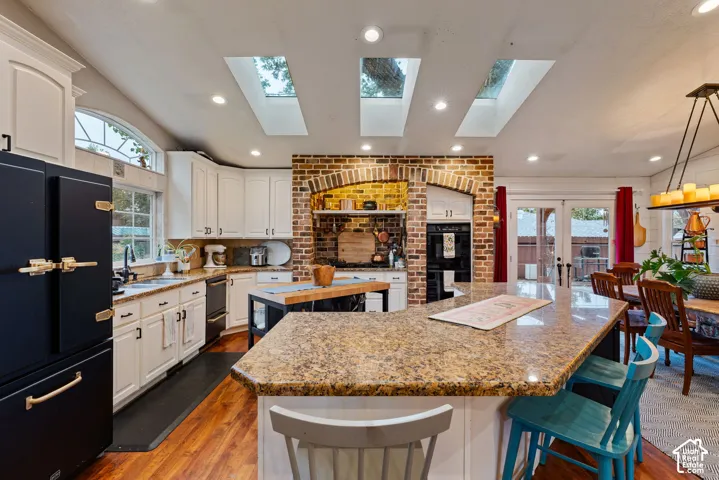 Kitchen featuring white cabinetry, stone countertops, central island, exposed brick arched alcove, and triple skylights over wood-finish flooring