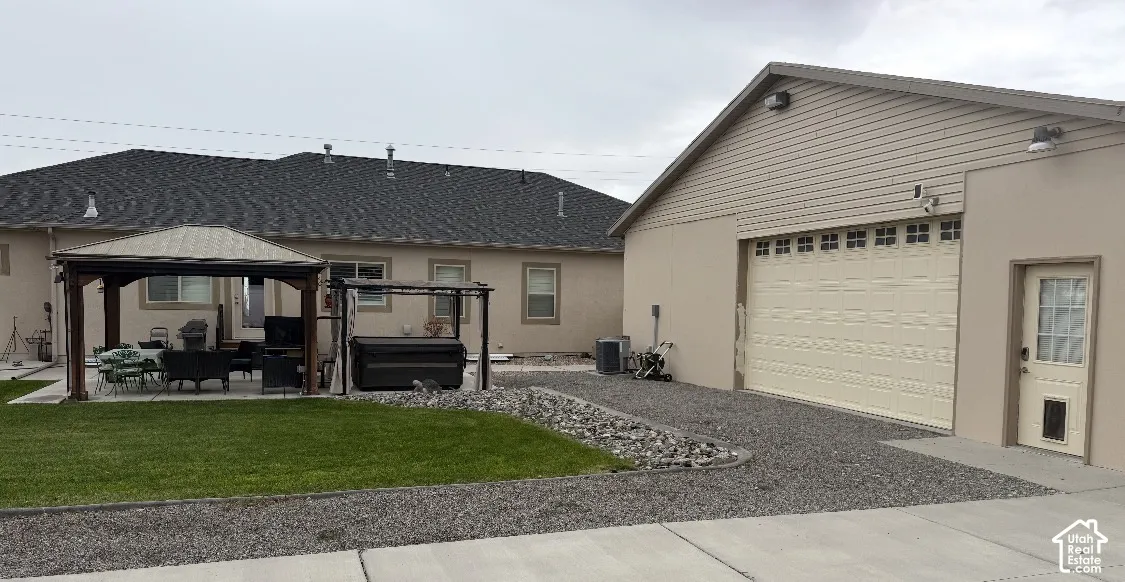 Stucco exterior featuring a detached garage, concrete patio, built-in hardtop gazebo, and manicured lawn