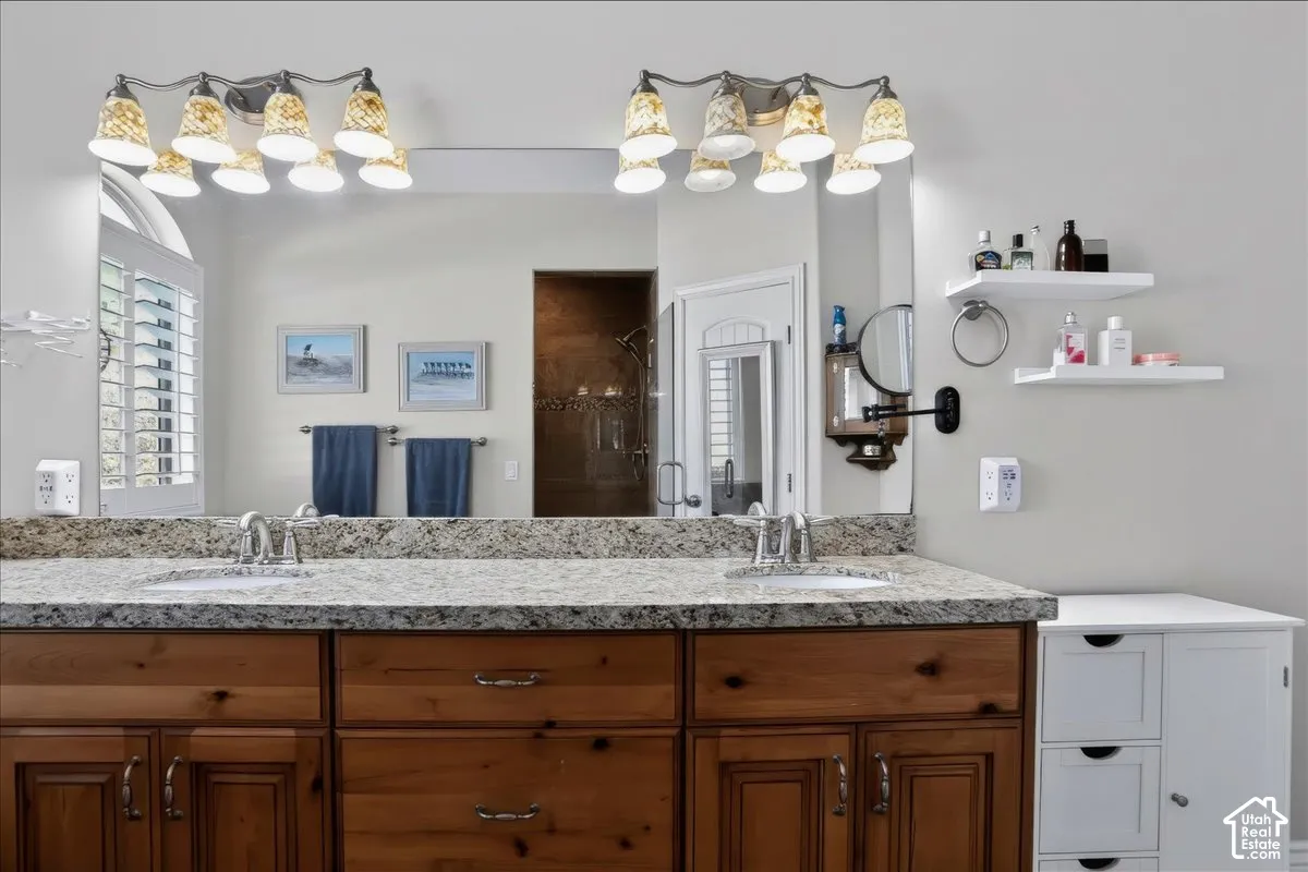 Dual-sink vanity featuring light-toned stone countertop, wood cabinetry, two oversized light fixtures with glass shades, and a full-width wall mirror