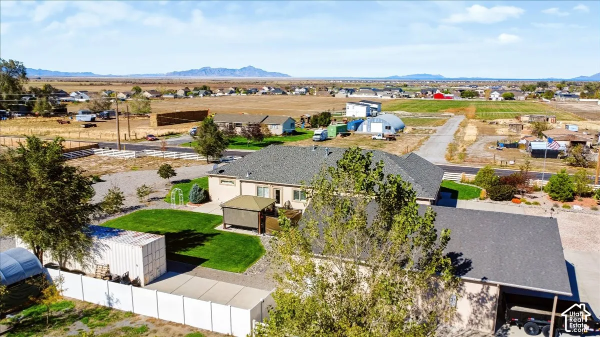 Aerial perspective of a property featuring a manicured lawn, white vinyl fencing, a detached storage structure, a covered hardscaped area, and mountainous horizons
