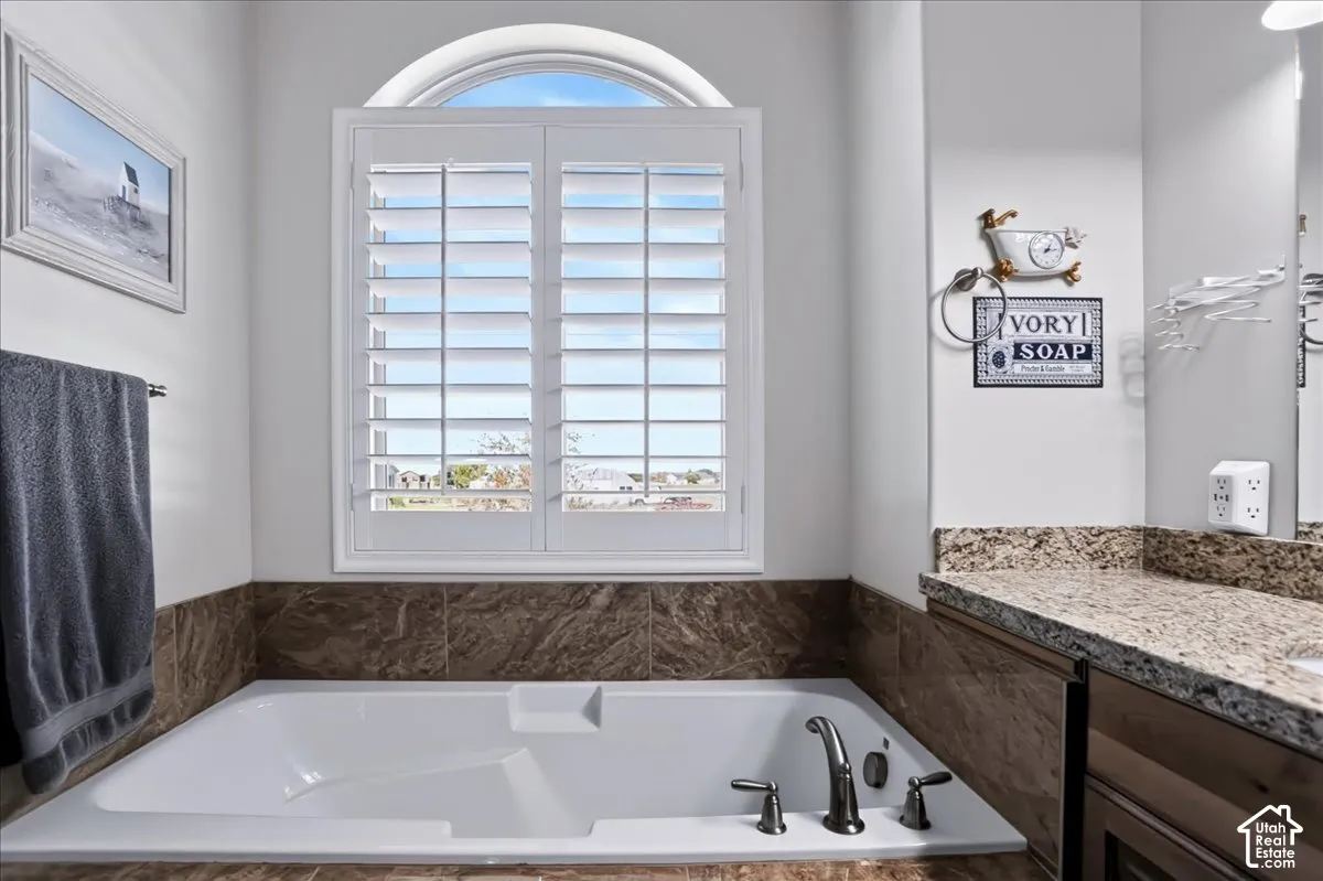 Built-in soaking tub with dark stone-finish surround, white plantation shutters, arched transom window, and stone-finish vanity countertop