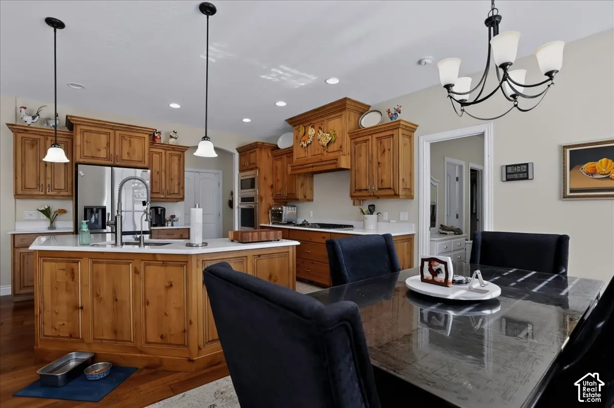 Kitchen featuring knotty wood cabinetry, light-toned countertops, a center island with a stainless steel sink, built-in double wall ovens, and pendant and chandelier lighting