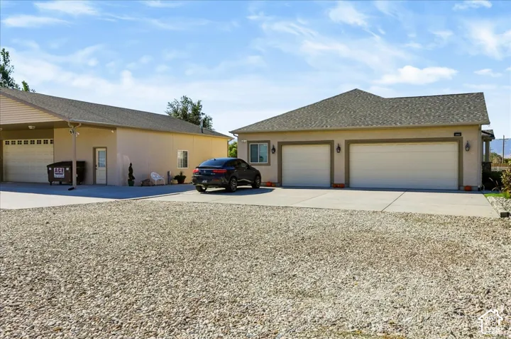 Stucco exterior featuring a two-car attached garage and an additional detached structure with a covered parking bay, concrete driveway, and gravel lot