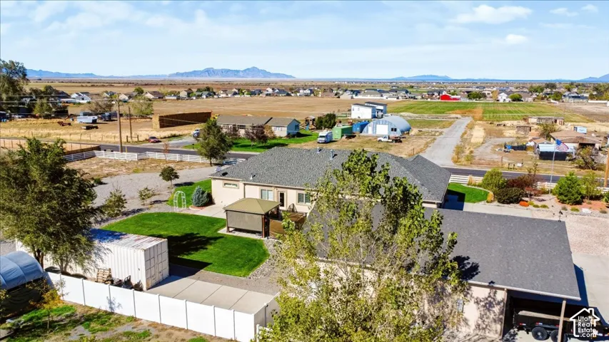 Aerial perspective of a property featuring a manicured lawn, white vinyl fencing, a detached storage structure, a covered hardscaped area, and mountainous horizons