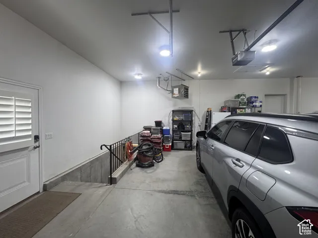 Finished garage featuring smooth concrete flooring, integrated ceiling lighting, a door with plantation-style shutters, and a metal stair railing