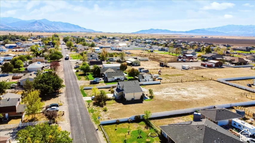 Aerial perspective featuring a multi-story residence with a pitched shingle roof, expansive open lot, white perimeter fencing, and a prominent mountain range backdrop